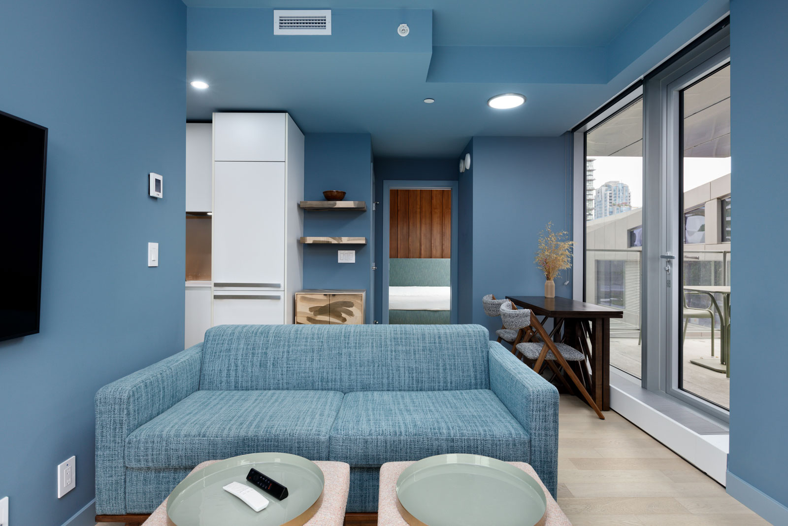 Modern living room with blue walls and couch, round coffee tables, and large glass door opening to a balcony. A small desk and chairs are in the corner, with a glimpse of a bedroom visible.