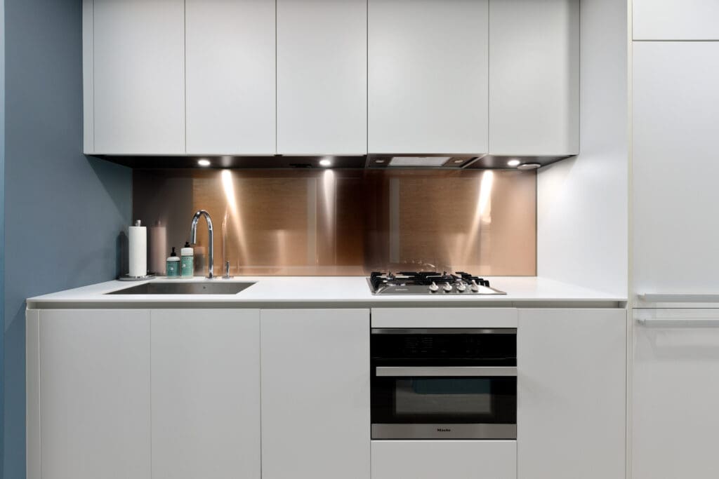 Modern kitchen with white cabinets, built-in gas stove, oven, and stainless steel backsplash. Sink on the left with a faucet and soap dispensers. Minimalist design with under-cabinet lighting.