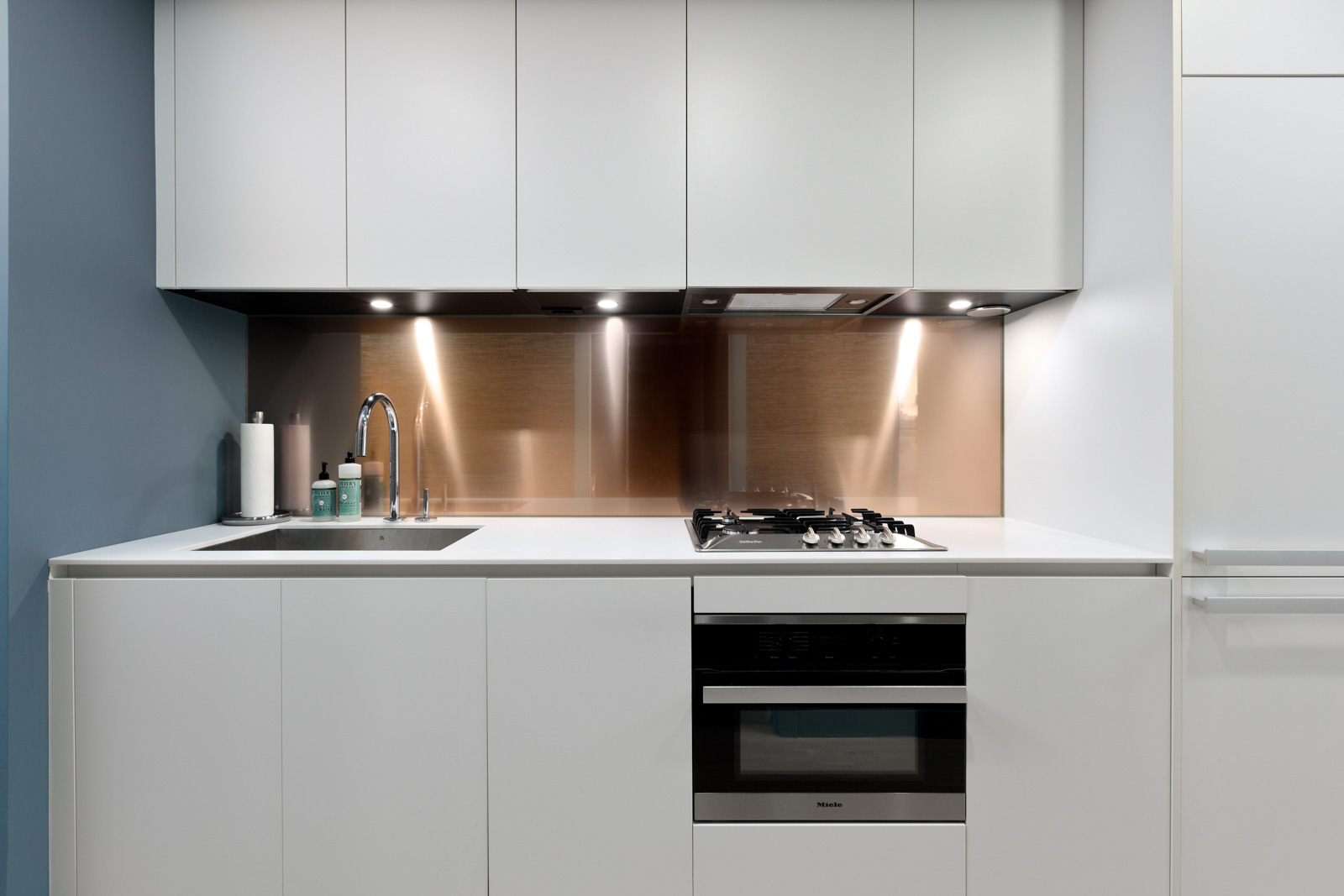 Modern kitchen with white cabinets, built-in gas stove, oven, and stainless steel backsplash. Sink on the left with a faucet and soap dispensers. Minimalist design with under-cabinet lighting.