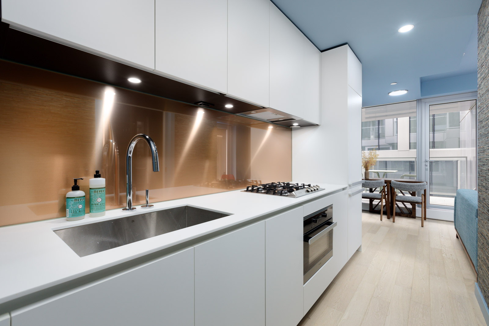 Modern kitchen with white cabinets, stainless steel sink, gas stovetop, and copper backsplash. Beyond, a dining area with a table and chairs is visible through large windows.