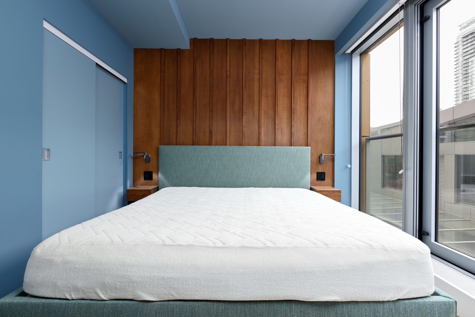 A modern bedroom features a blue-themed decor with a large bed, wooden accent wall, and floor-to-ceiling windows overlooking a cityscape.