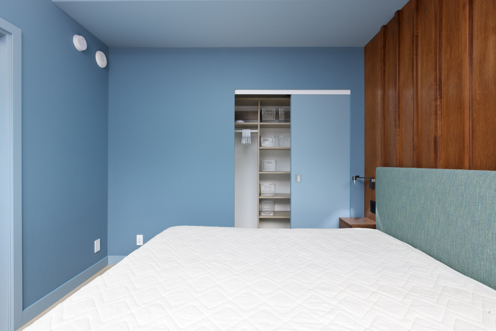 Minimalist bedroom with blue walls, a closet door slightly open showing shelves, and a bed with a textured white bedspread. Wood paneling accents one wall.