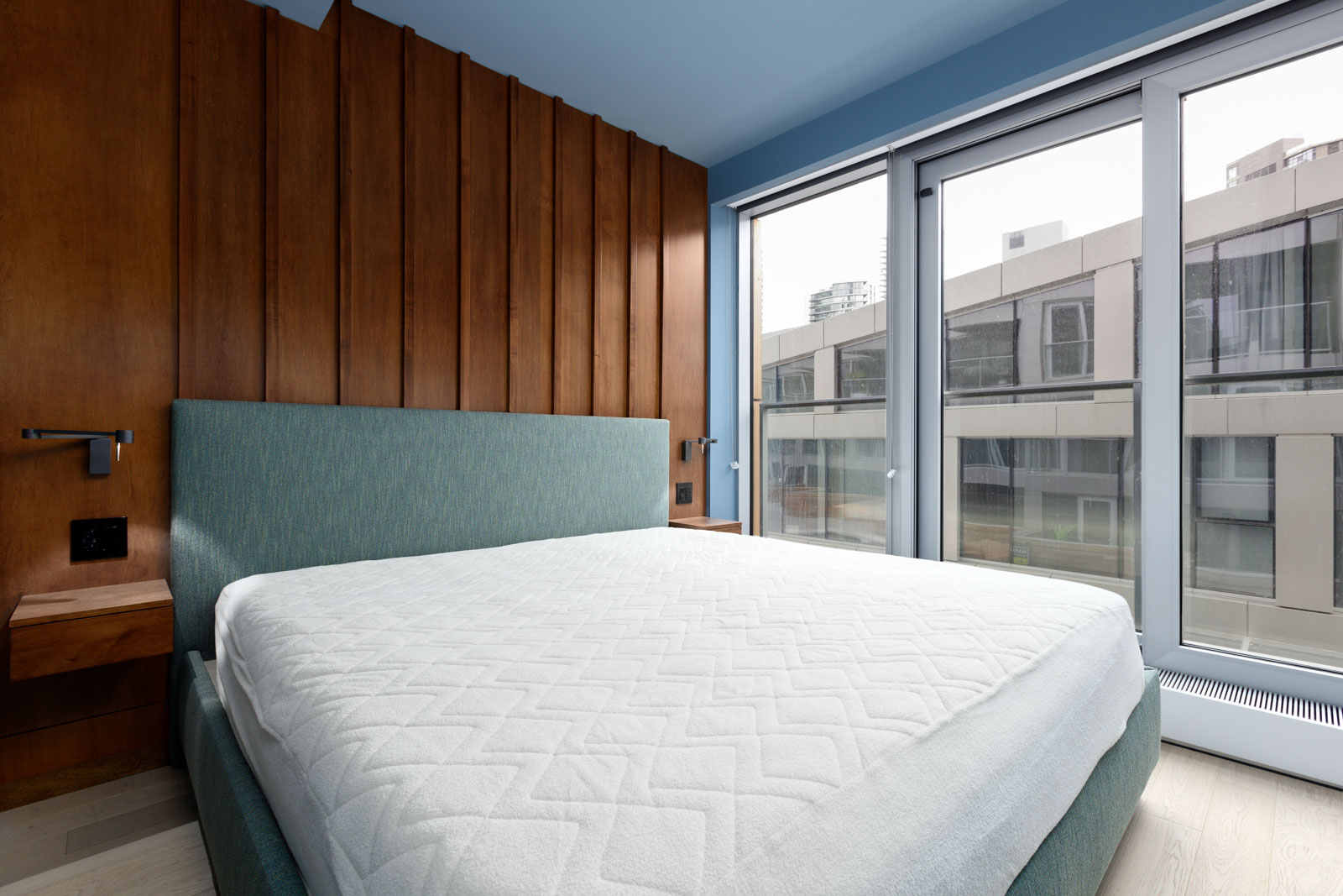 Modern bedroom with a teal upholstered bed, wooden wall panels, and large windows overlooking an urban building. Natural light fills the room.