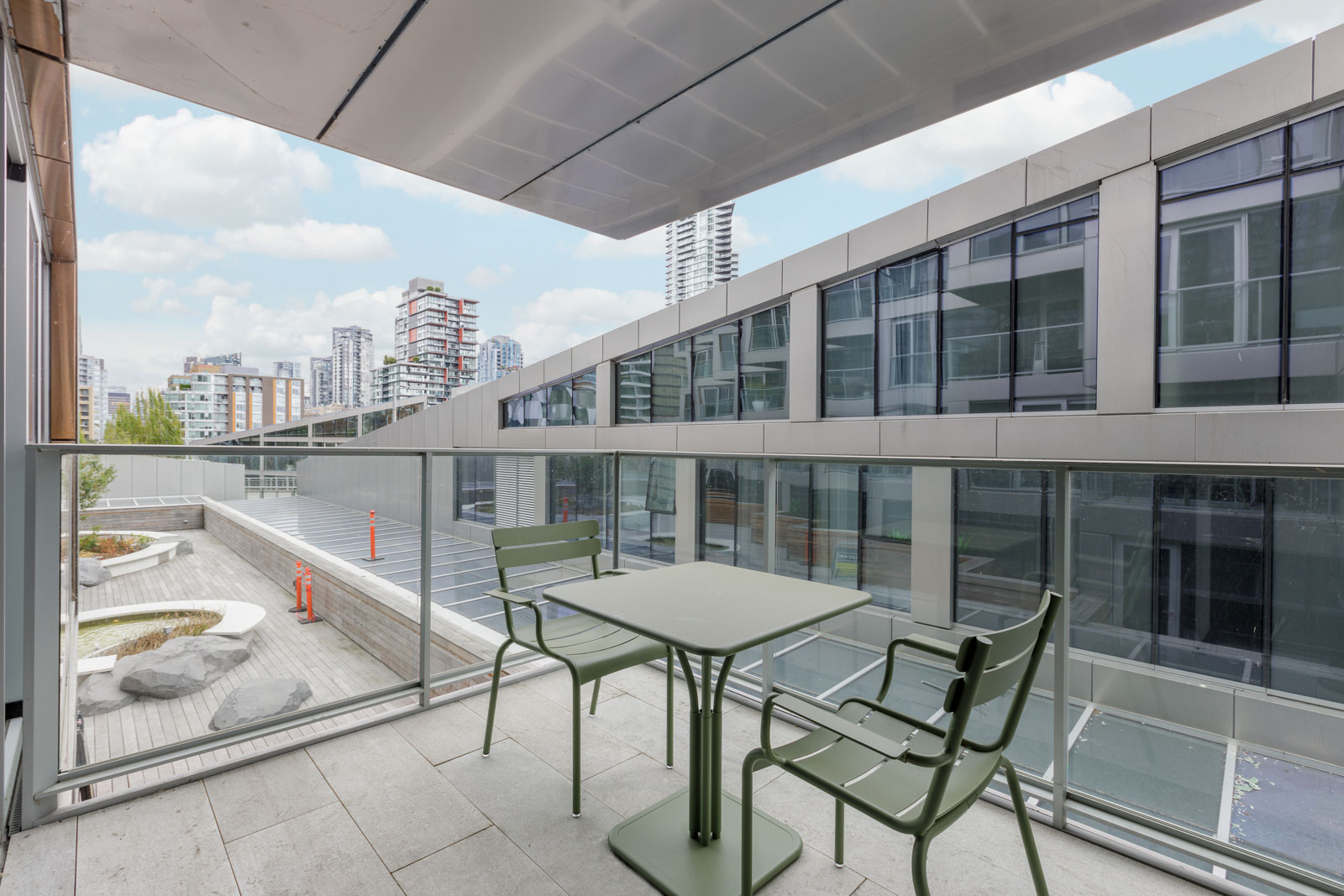 Modern balcony with a green table and two chairs overlooking a courtyard and contemporary buildings, under a cloudy sky.