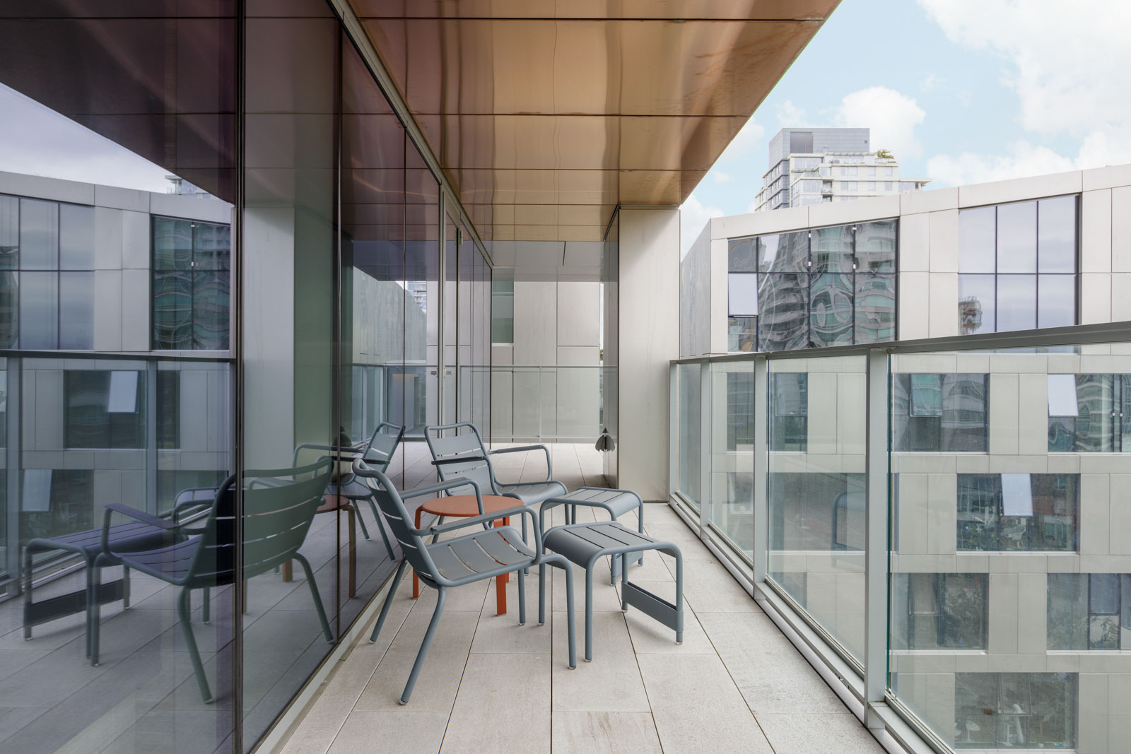 Modern balcony with glass railing, featuring three metal chairs and a table. High-rise buildings are reflected in the glass, with a clear sky overhead.