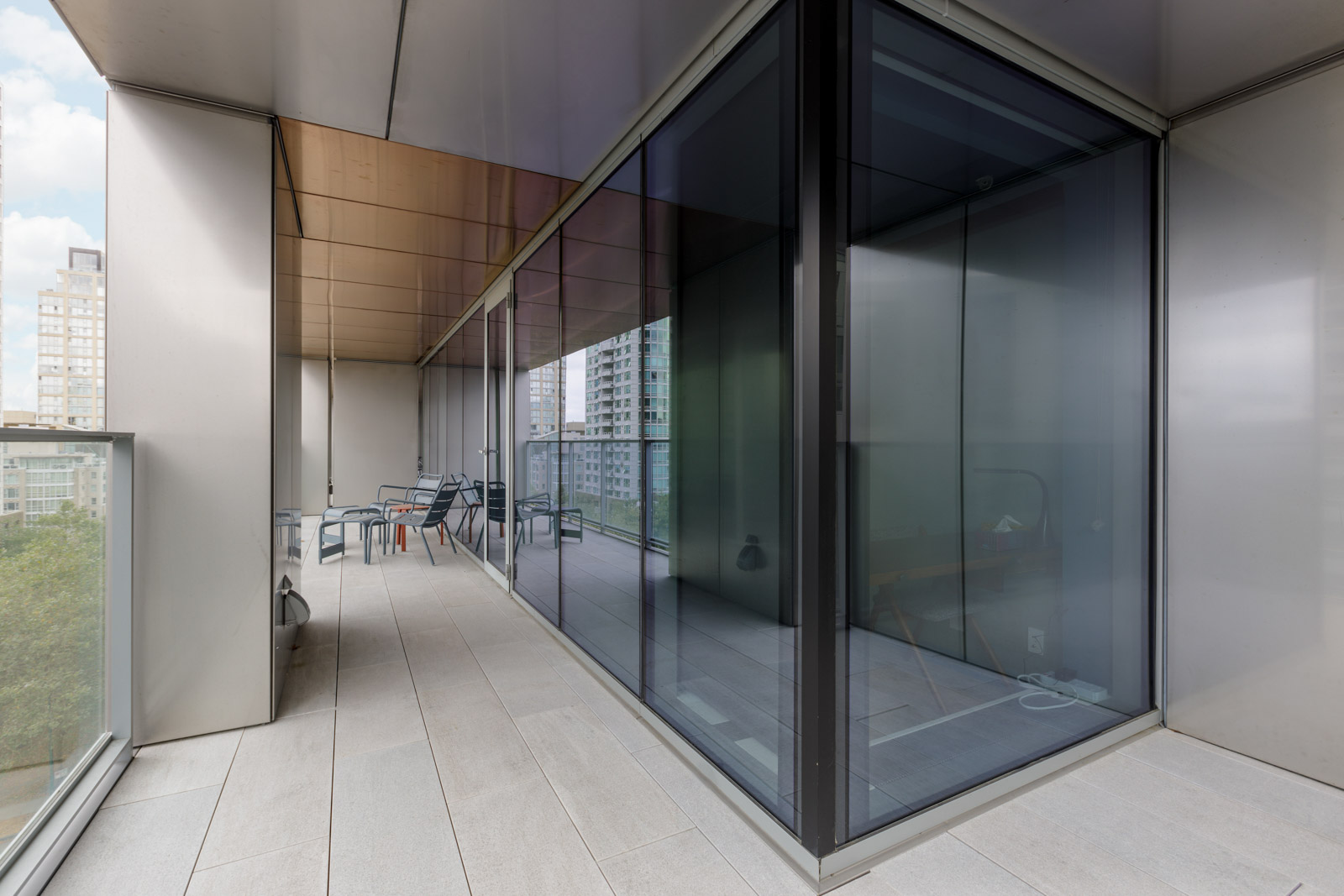 A spacious balcony with modern glass walls and outdoor furniture, overlooking a cityscape with tall buildings.