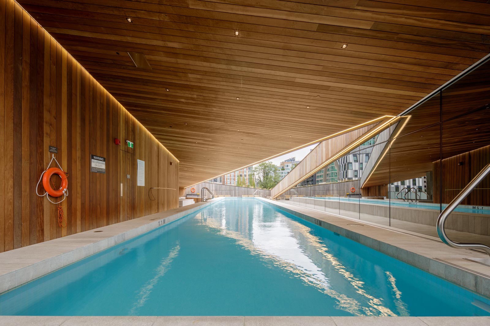 Indoor swimming pool with wooden ceiling and walls, reflecting in a large mirror. Safety equipment and a modern building view are visible.