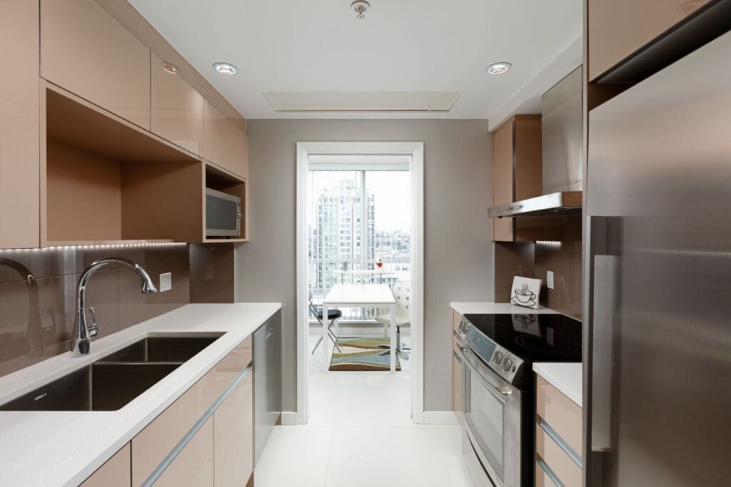 Modern kitchen with beige cabinetry, stainless steel appliances, and a white countertop. A window at the end of the kitchen shows a city view.