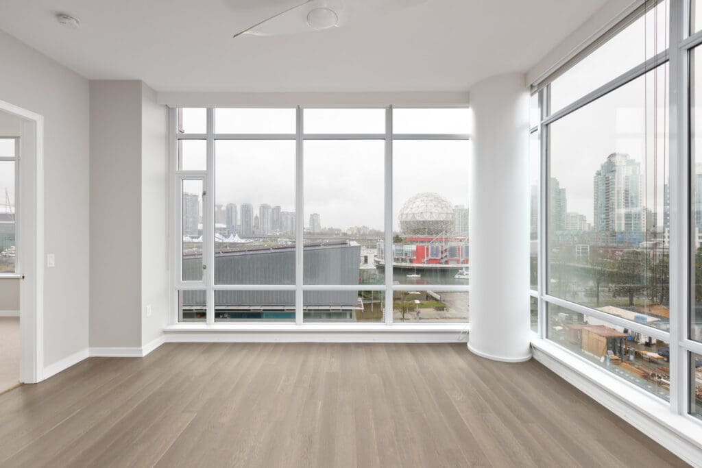 Spacious corner room with large windows showcasing a city skyline and waterfront. The room has wooden floors and neutral walls.