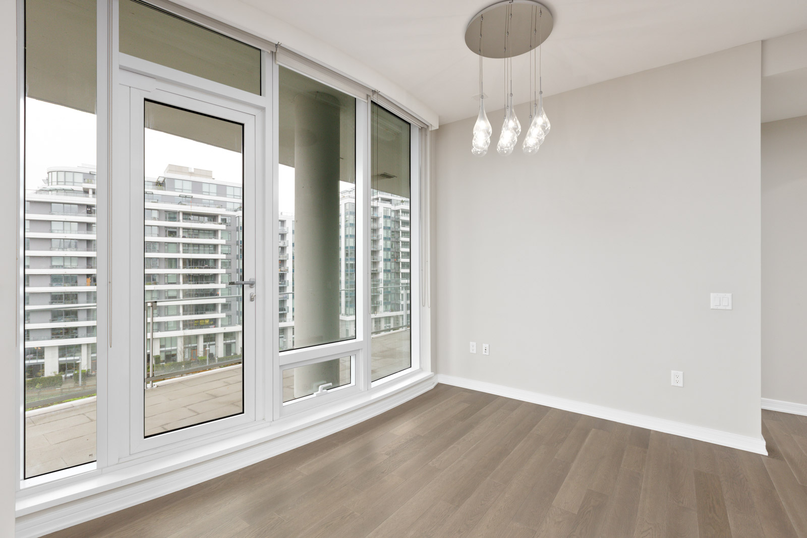 Empty room with wooden flooring, large windows, and a ceiling light fixture. The windows overlook modern apartment buildings.