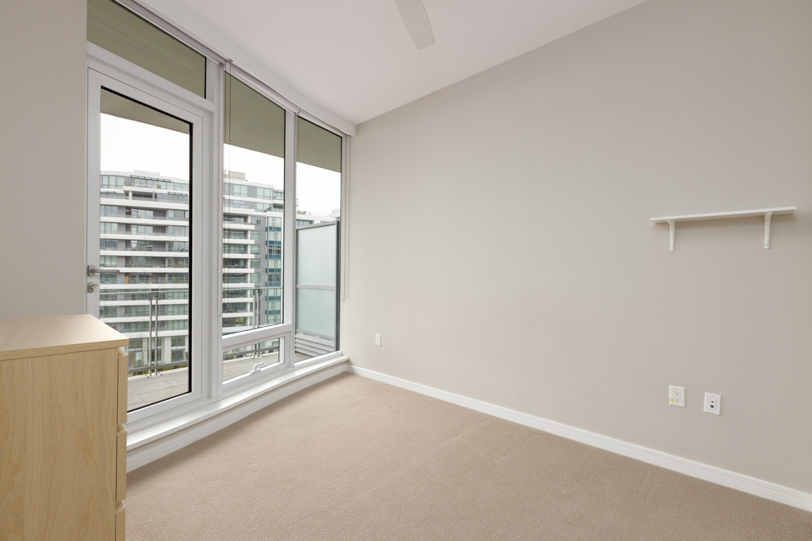 Empty room with beige carpet, white walls, and large windows with a view of modern buildings. A small wooden cabinet and a wall shelf are present.