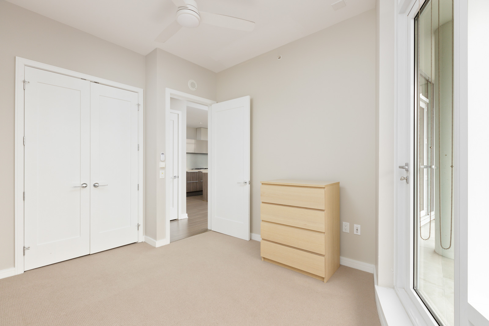 An empty room with a beige carpet, white walls, a ceiling fan, a light wood dresser, double white doors, and a doorway leading to a kitchen area.
