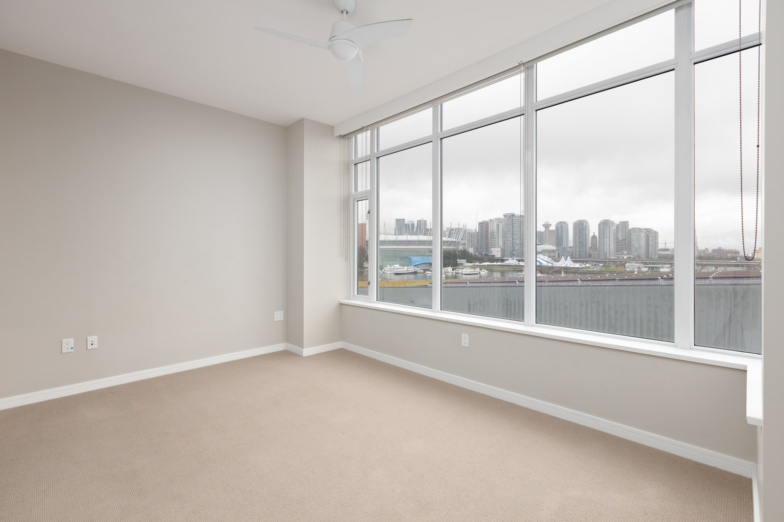Empty room with beige carpet and large windows showing a city skyline. Walls are light gray with a ceiling fan present.