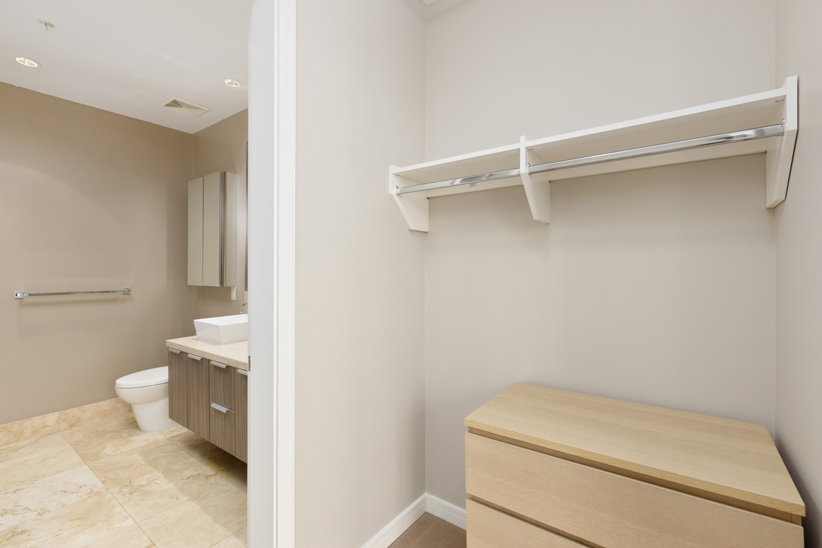 Minimalist bathroom and closet with a toilet, vanity, towel rack, shelving, and dresser. Cream and beige tones dominate the decor.