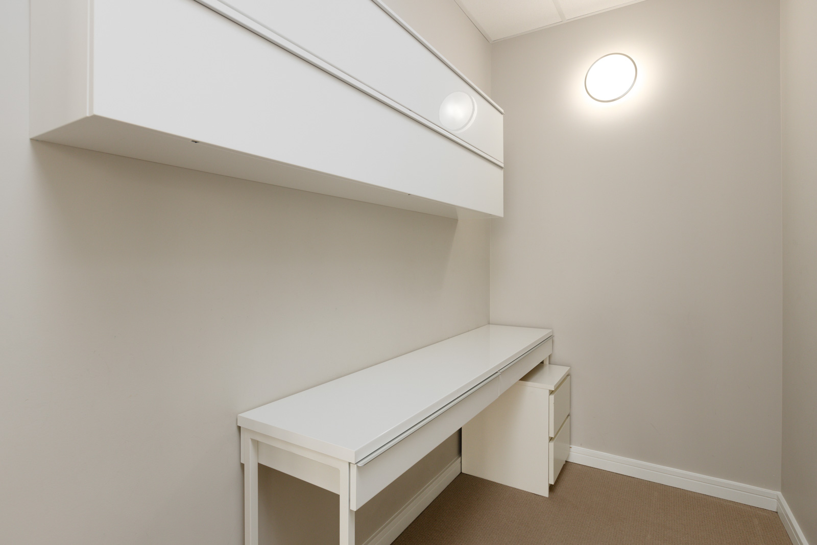 Minimalist white office space with a long desk, wall-mounted storage, and a small drawer unit. A round light fixture is on the wall.