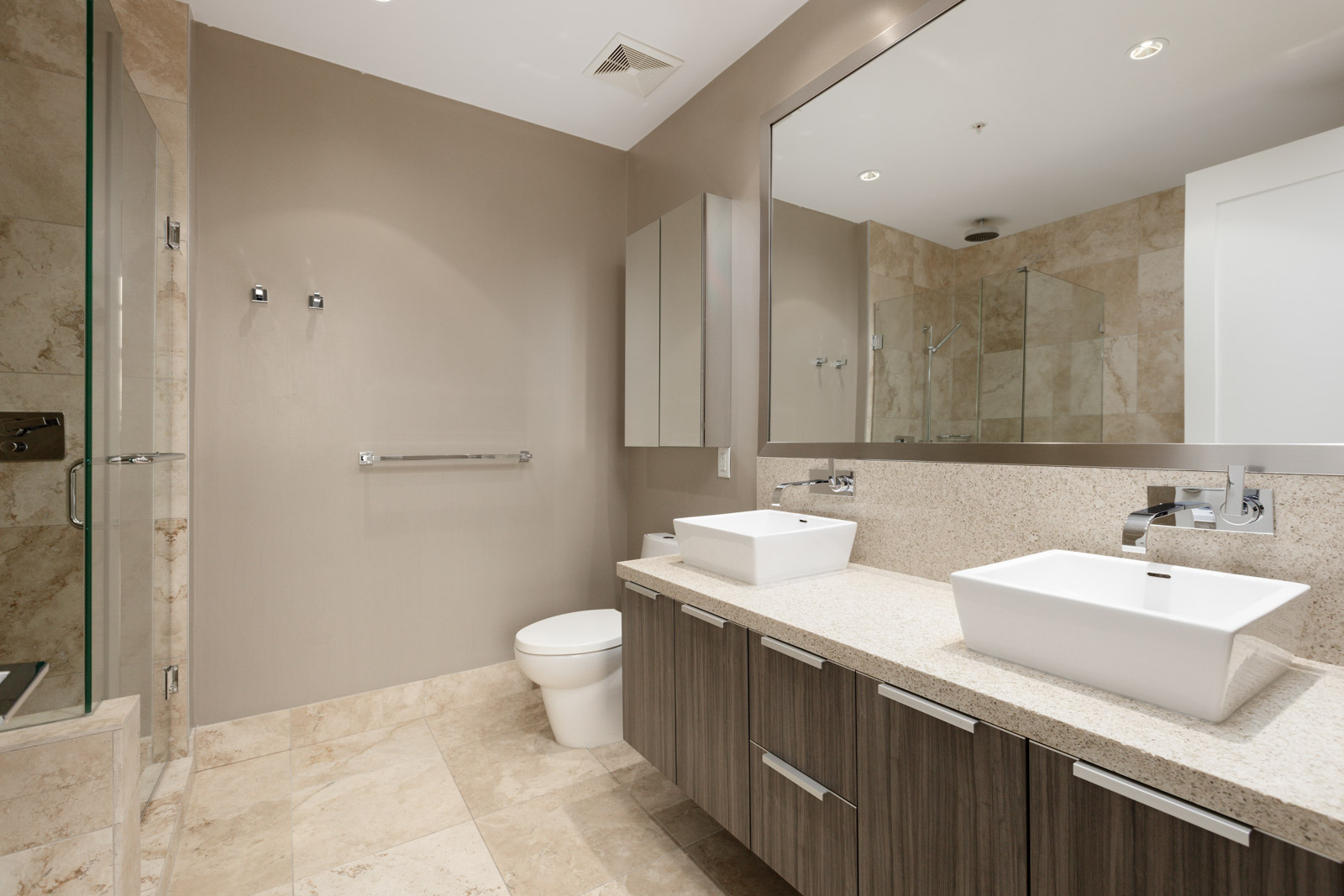Modern bathroom with two vessel sinks on a wooden vanity, large mirror, beige walls, tiled floor, and a glass-enclosed shower.