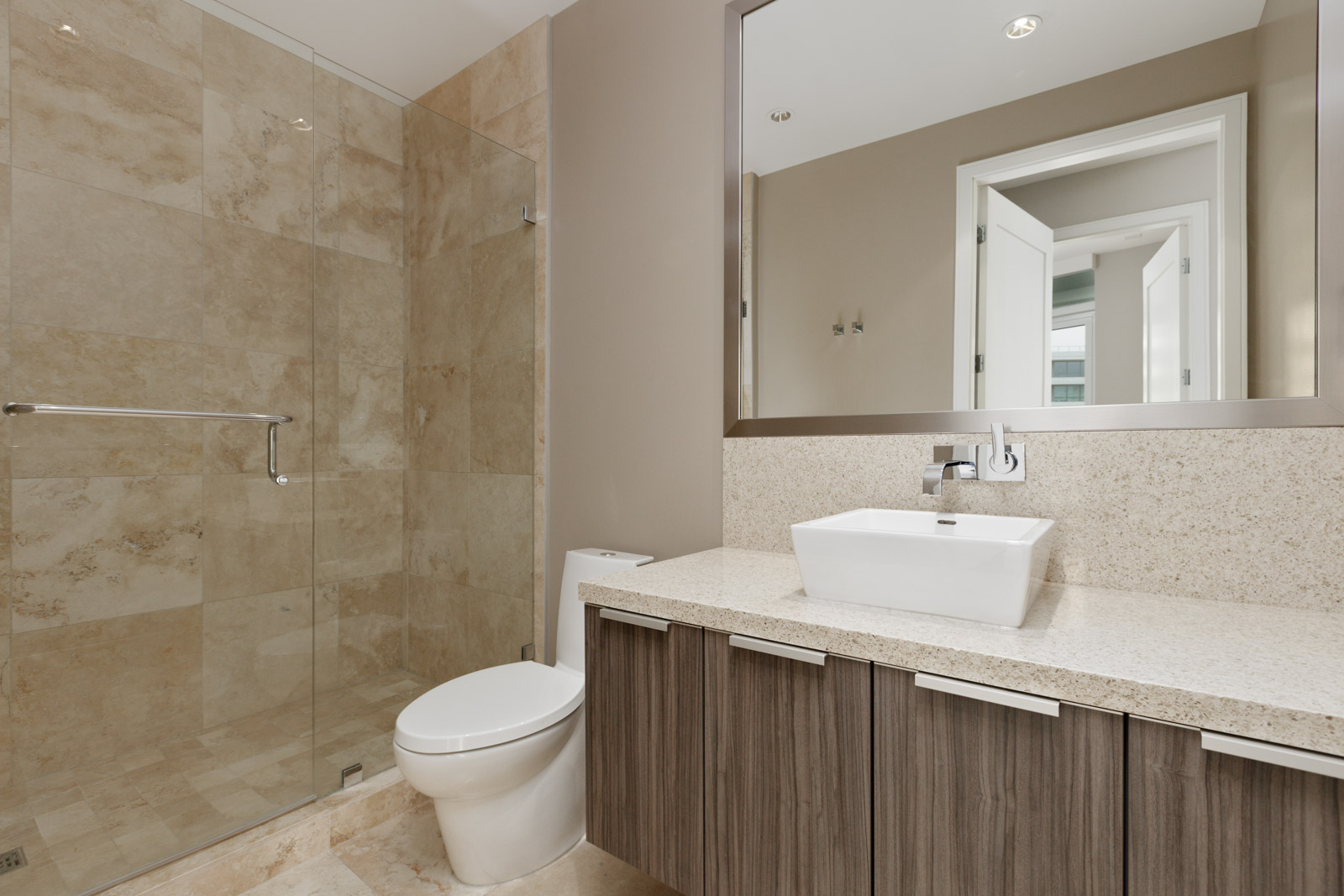 Modern bathroom with a glass-enclosed shower, beige tile, a white toilet, and a wooden vanity with a white rectangular sink and large mirror.