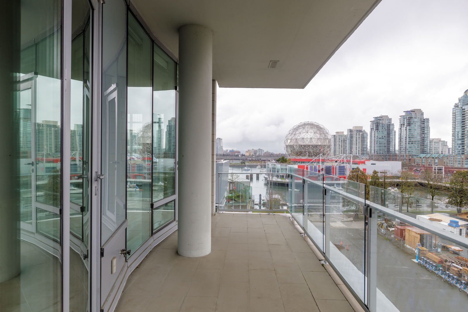 A glass balcony with a city skyline view, including a dome structure and high-rise buildings in the background. Overcast sky visible.