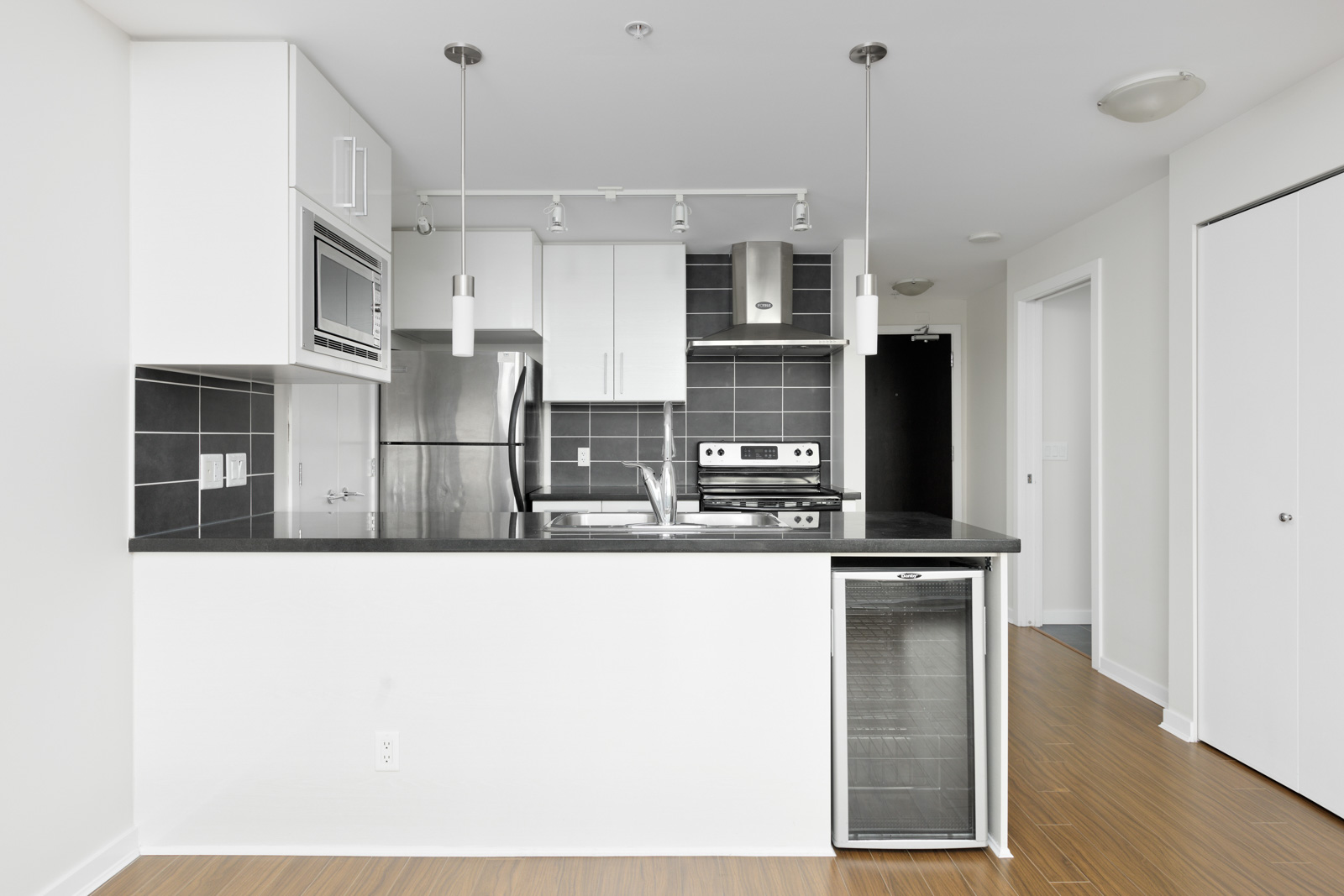 Modern kitchen with stainless steel appliances, black tile backsplash, and wooden floors. Features a white counter and suspended lights.
