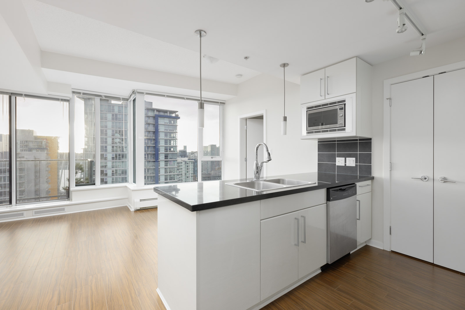 Modern kitchen with a black countertop and stainless steel appliances, including a microwave and dishwasher. Large windows offer a city view, and light wood floors complete the space.
