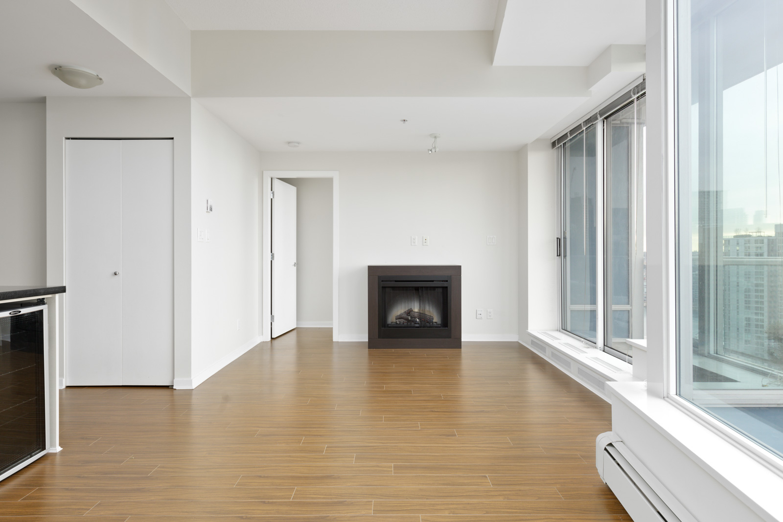 A modern, empty living room with hardwood floors, a fireplace, large windows, and white walls in a high-rise apartment.