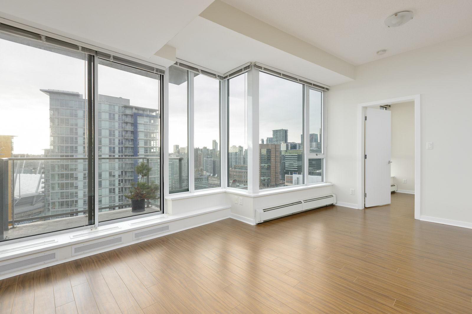 A bright, empty room with wooden flooring features large windows offering a cityscape view. A small plant sits on the windowsill. A doorway leads to another room.