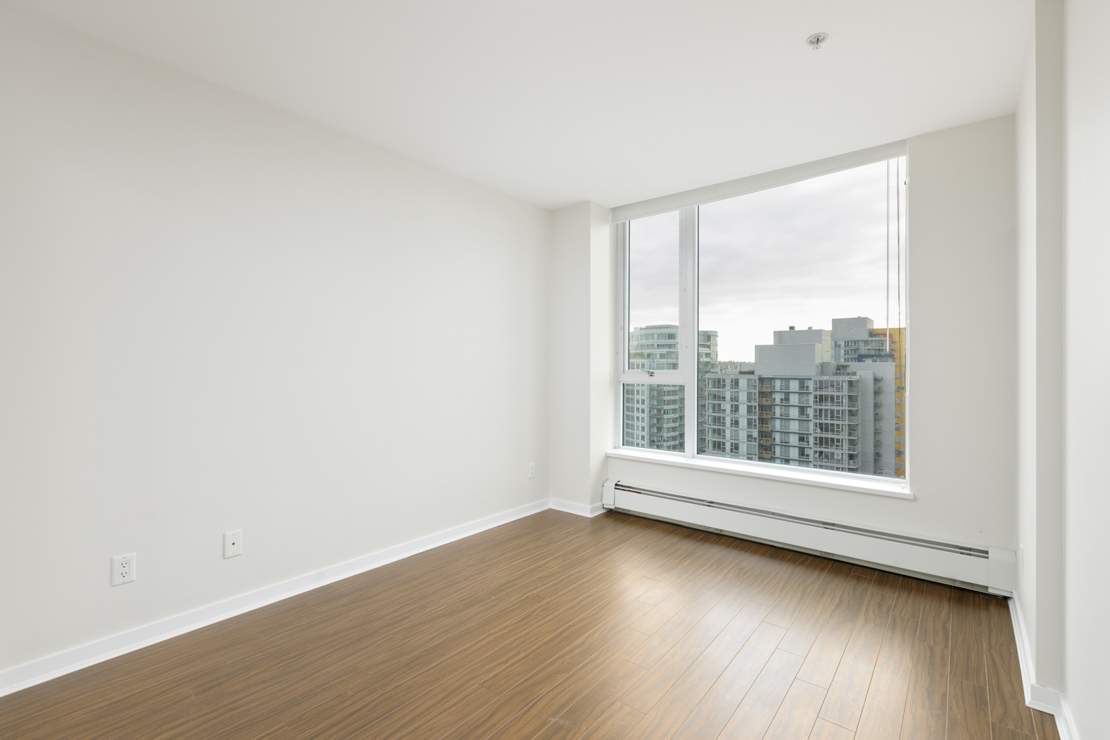 Empty room with large window, white walls, and wood flooring, overlooking city buildings.
