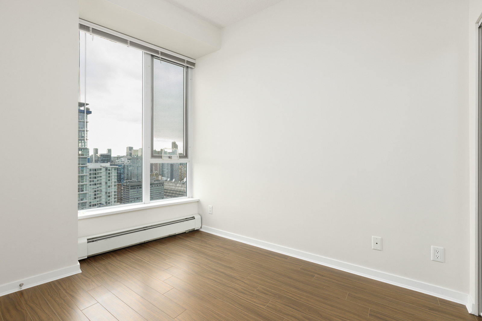 Empty room with light wood flooring, large window, and white walls, overlooking a cityscape with tall buildings.