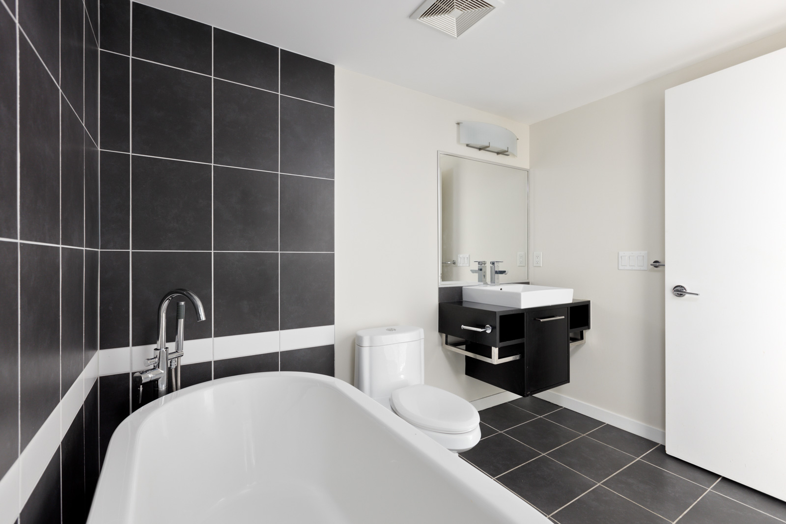 Modern bathroom with a white bathtub, black and white tiled walls, a wall-mounted sink with a mirror above, and a toilet. The room features a white door and minimalist design.