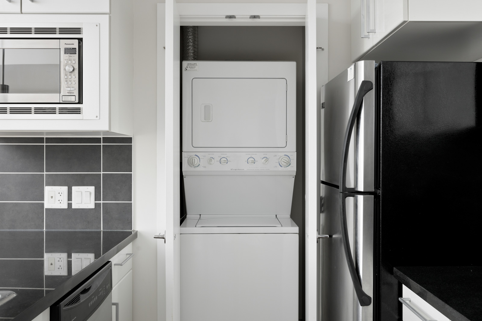 Compact kitchen space featuring a stainless steel refrigerator and a built-in microwave with a stacked washer and dryer unit neatly enclosed in a closet.