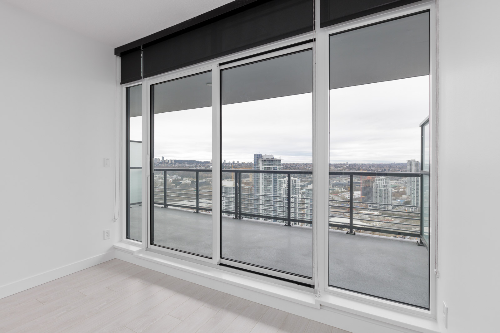 Empty room with light wood flooring and large glass sliding doors leading to a balcony. The view shows a cityscape with tall buildings and overcast sky.