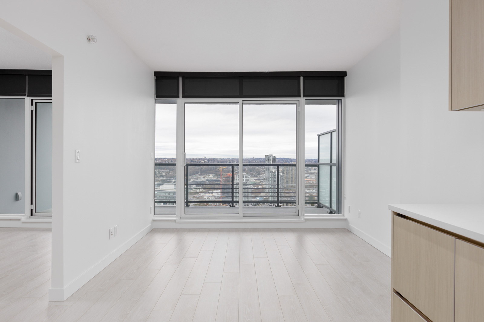 Modern empty room with large windows and a balcony overlooking a cityscape. Light wooden flooring and white walls enhance the bright, open space.