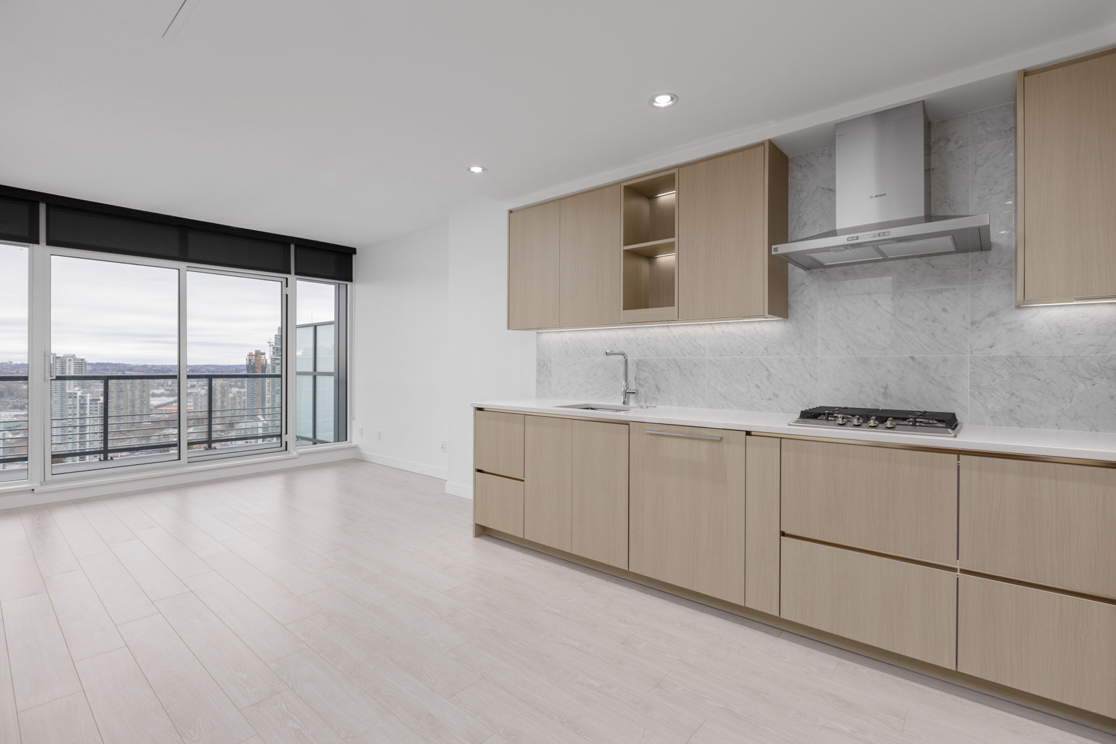 Modern kitchen with light wood cabinets, marble backsplash, and built-in stovetop. Large windows offer a view of a cityscape and balcony. Light wood flooring throughout.