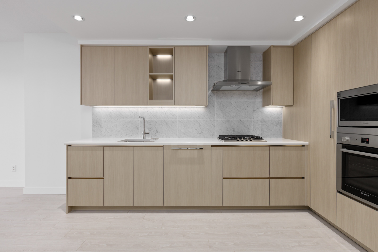 Modern kitchen with light wood cabinets, marble backsplash, built-in oven, gas stove, and stainless steel range hood, featuring recessed lighting and a light wood floor.