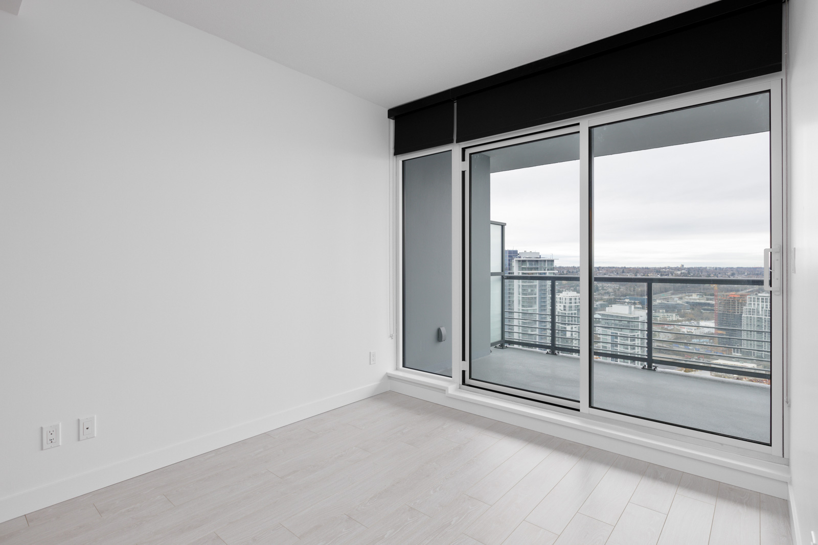Empty room with light wood flooring and large sliding glass doors leading to a balcony with a cityscape view.