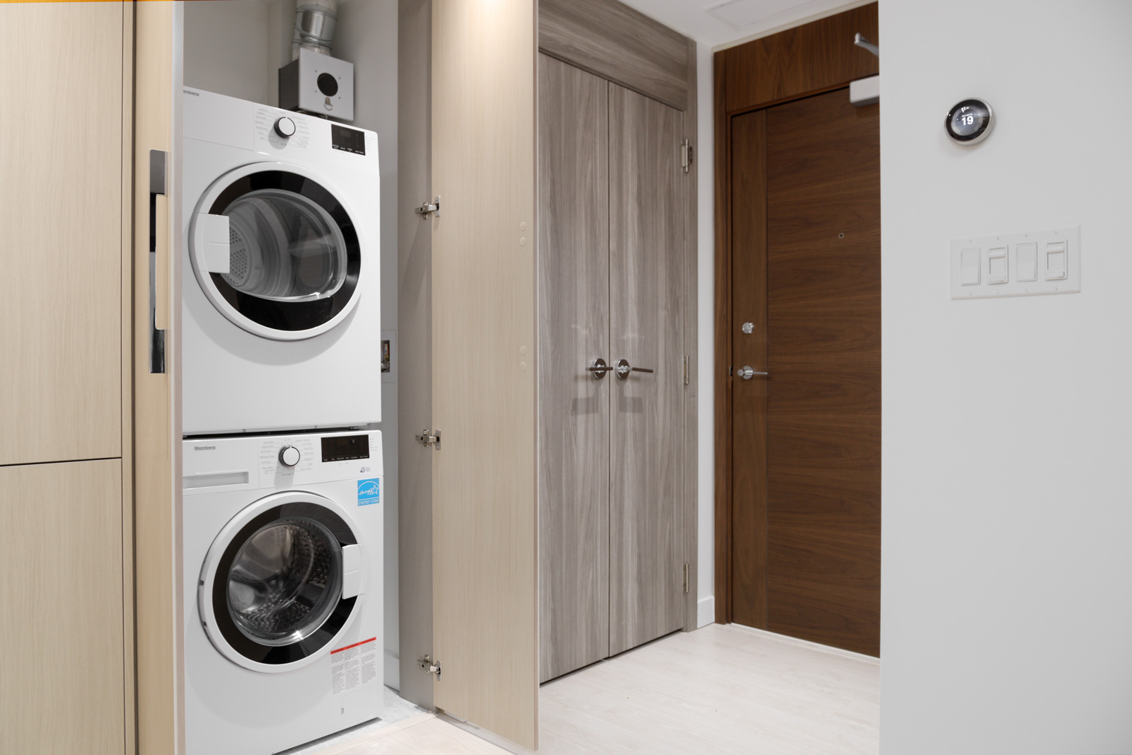 Stacked white washer and dryer in a small laundry area with wooden doors, adjacent to a brown entrance door.
