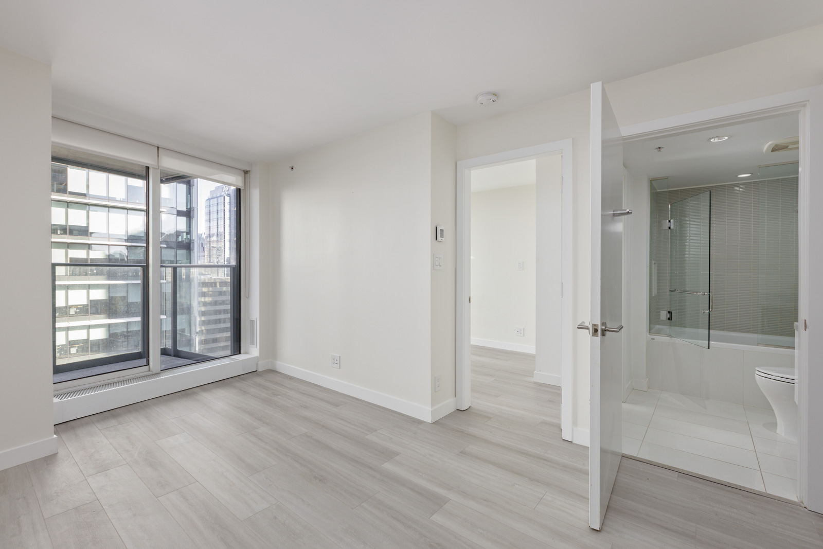 A modern, unfurnished apartment with large windows, light wood flooring, an open door leading to a bedroom, and a door leading to a bathroom with a bathtub and glass shower enclosure.