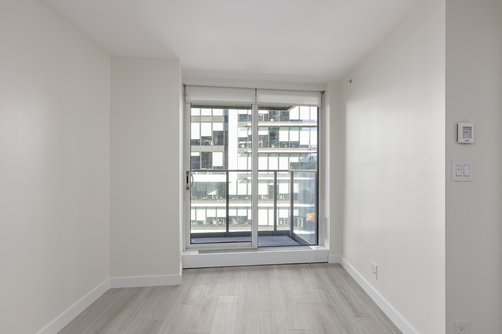 A room with white walls and light wooden flooring features a large floor-to-ceiling window with a sliding door, opening to a small balcony and view of a modern building exterior.