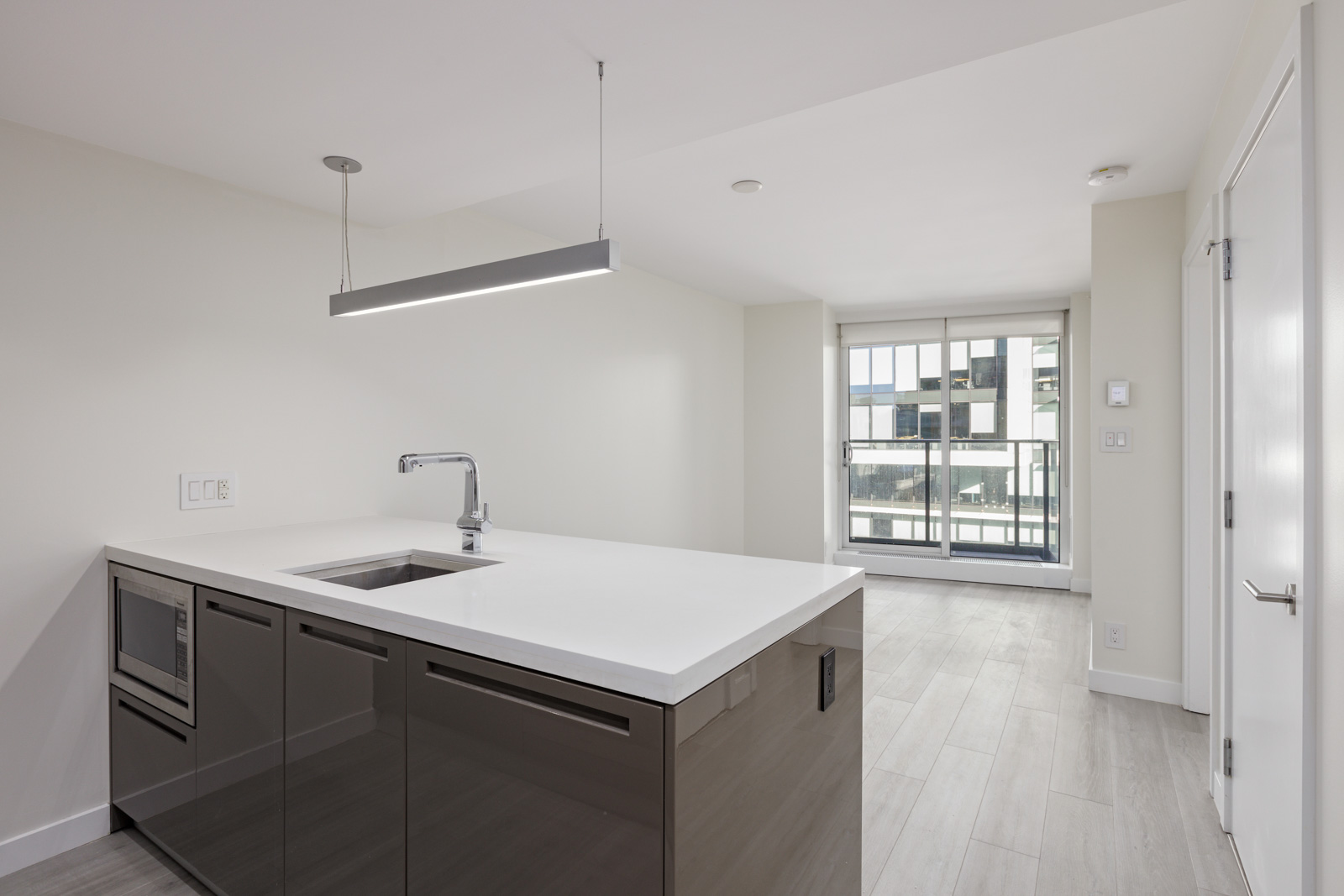 Modern kitchen with a sleek island featuring a sink and built-in appliances, minimalist white countertops, light wood flooring, and large windows in the background allowing natural light.