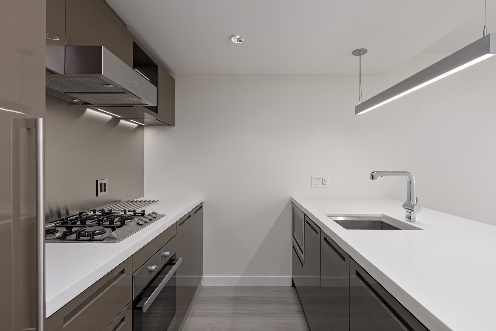 Modern kitchen with stainless steel gas stovetop, oven, and faucet. Sleek white countertops and grey cabinets. Minimalist design with overhead lighting.