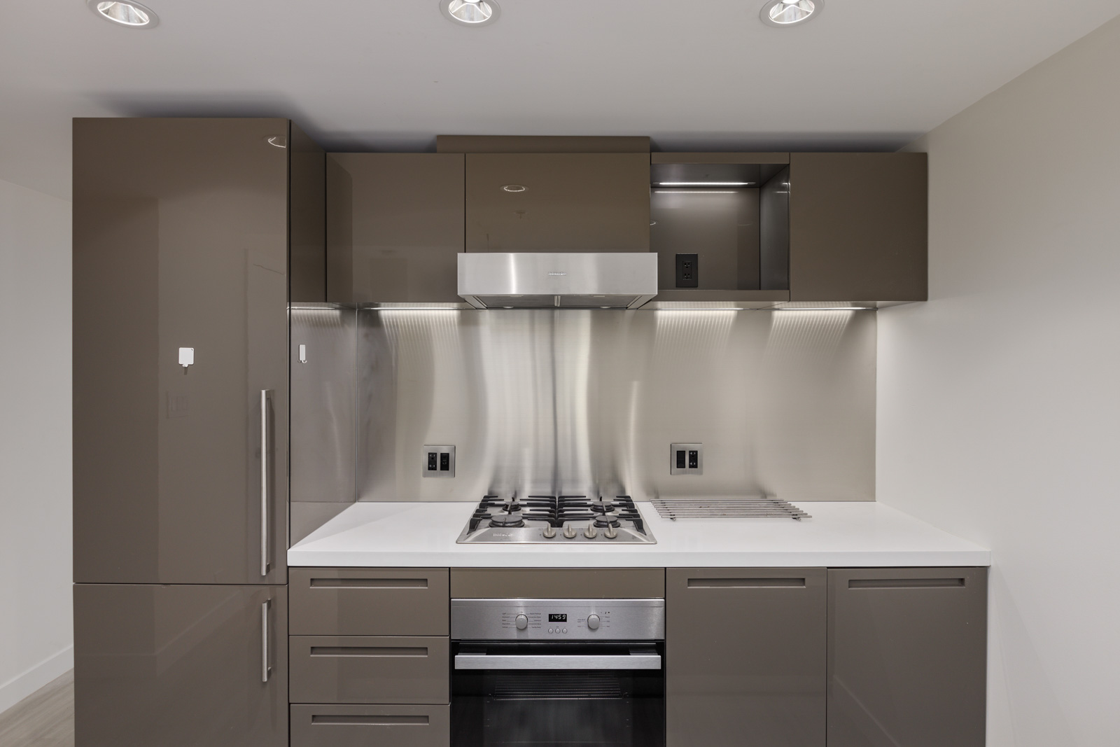 Modern kitchen with sleek brown cabinets, stainless steel gas stove and range hood, integrated refrigerator, and white countertops. Minimalist design with clean lines and under-cabinet lighting.
