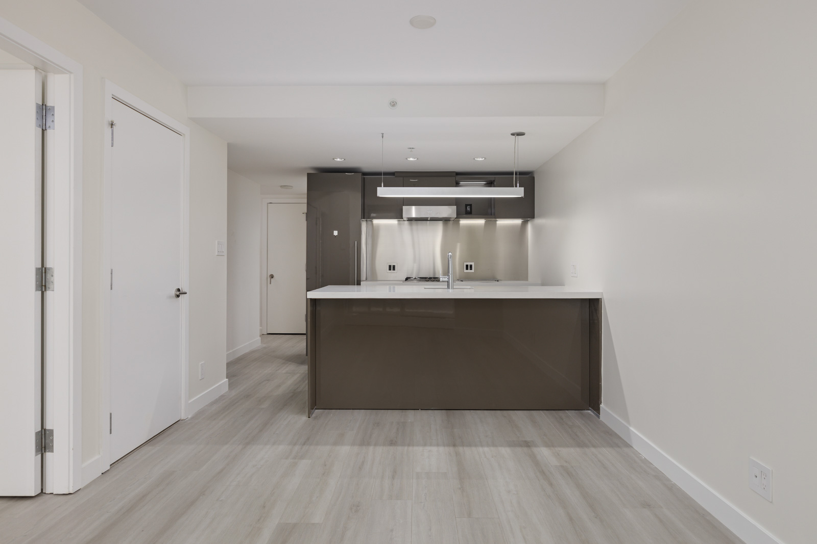 A modern kitchen with a glossy island, stainless steel appliances, pendant lights, and hardwood flooring. White walls and ceiling complement the sleek design. Door and hallway visible on the left.