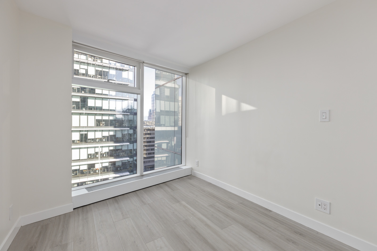 A minimalist, unfurnished room with light wood flooring, large windows, and a view of tall, modern buildings outside. The walls are painted white.