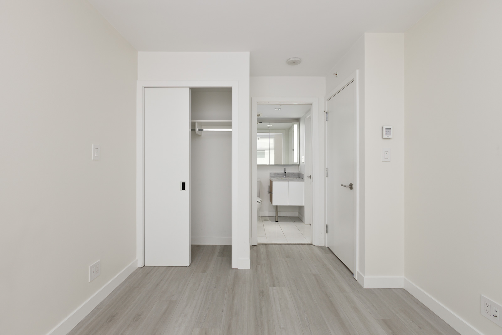 A minimalist room with light gray laminate flooring, a sliding door closet, and an open door leading to a small bathroom with a sink and mirror.