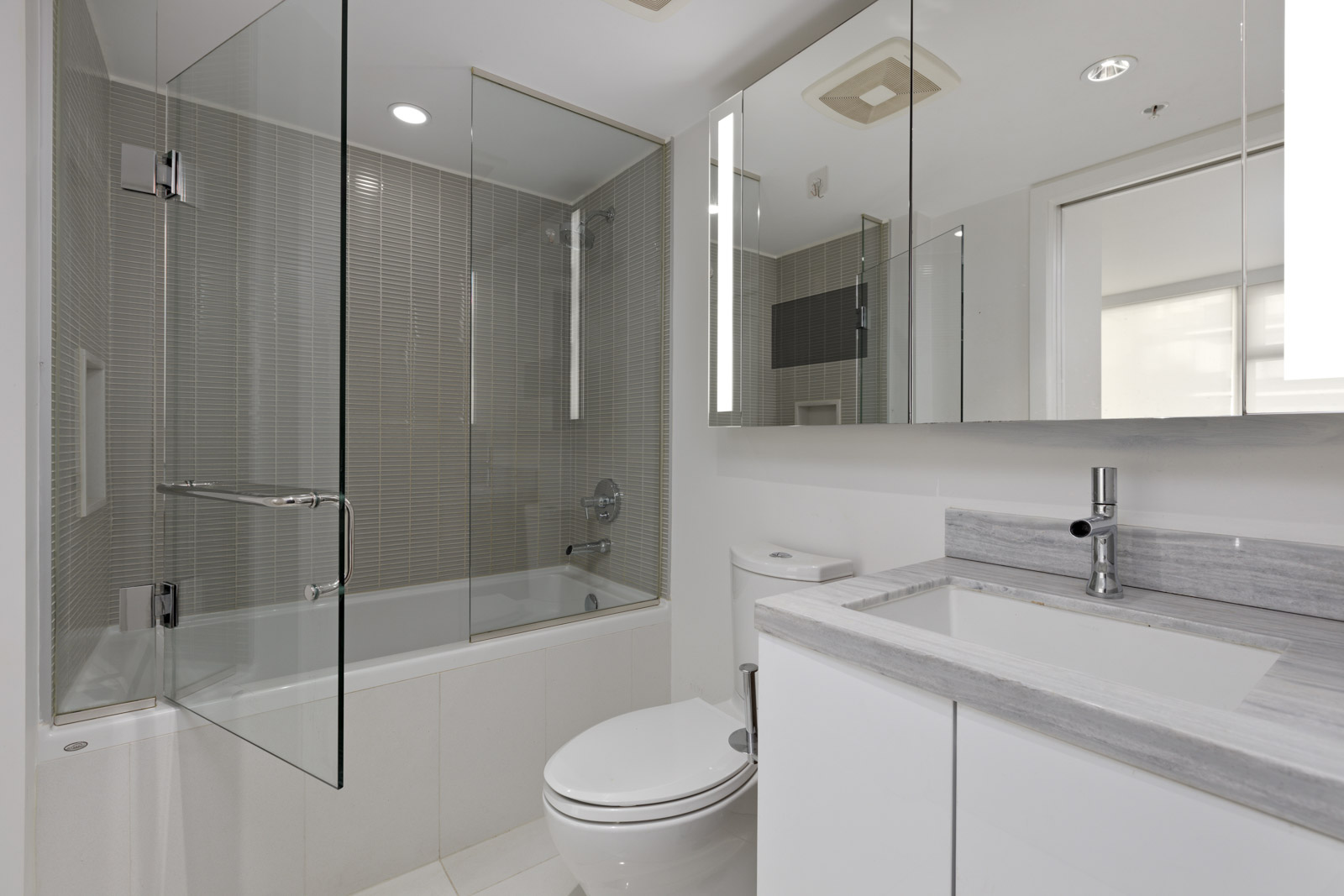 Modern bathroom with a glass-enclosed shower and bathtub, white toilet, sink with a gray countertop, mirrored cabinets, and neutral-colored tiles.