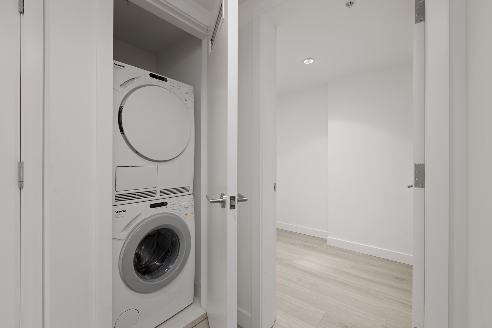 Stacked washer and dryer in a white laundry closet with doors slightly ajar, adjacent to a light-colored empty room with wood flooring.