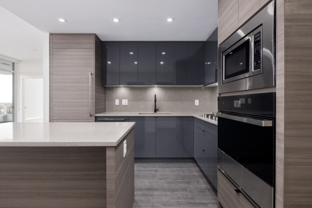 Modern kitchen with gray cabinets, stainless steel appliances, and an island. Black faucet, under-cabinet lighting, and wooden floor.