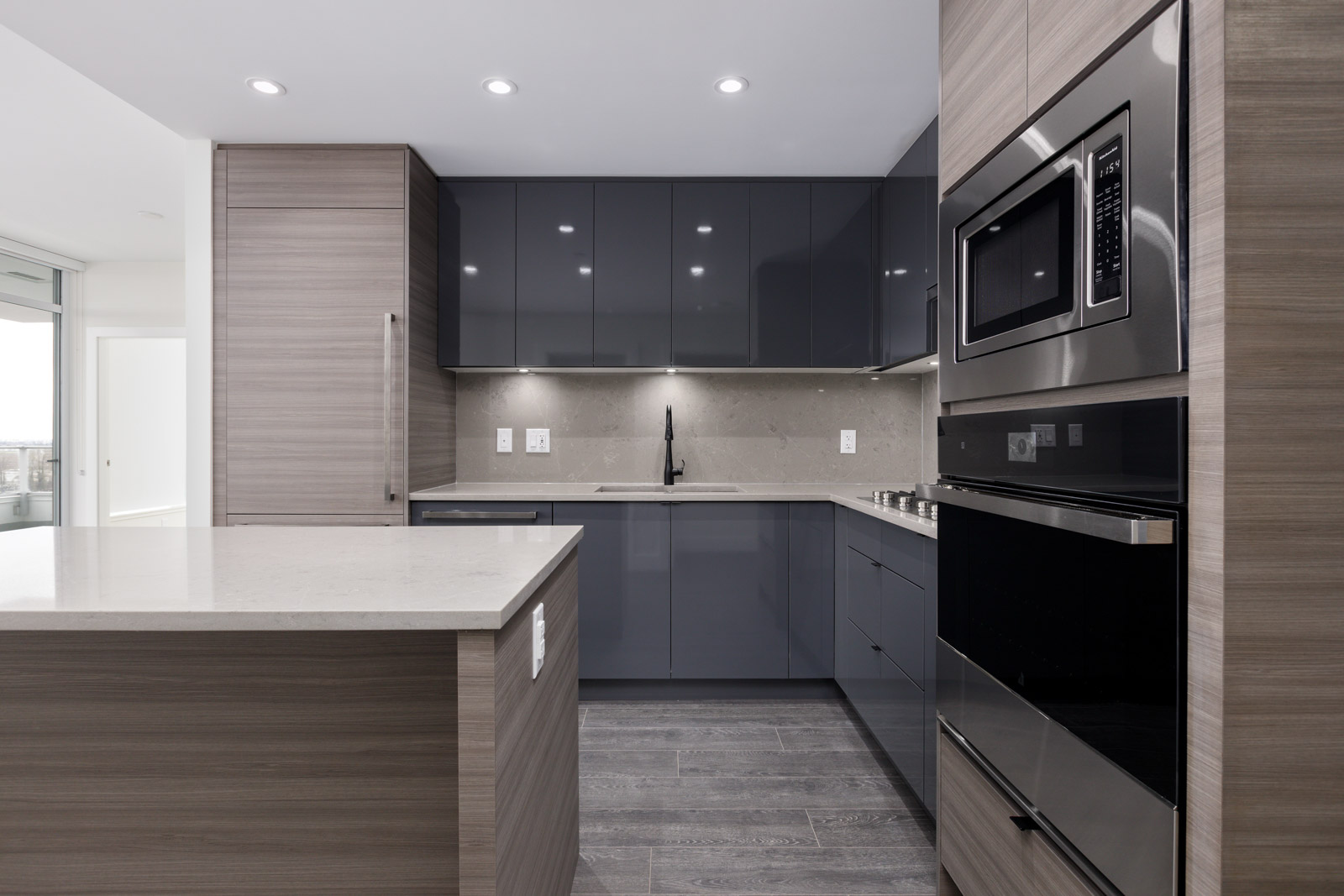 Modern kitchen with gray cabinets, stainless steel appliances, and an island. Black faucet, under-cabinet lighting, and wooden floor.
