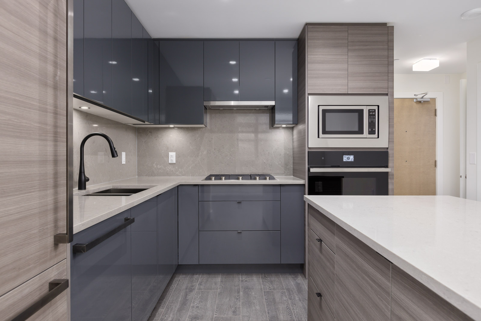 Modern kitchen with gray cabinets, stainless steel appliances, and a black faucet. Smooth countertops and wood-patterned flooring complete the sleek design.