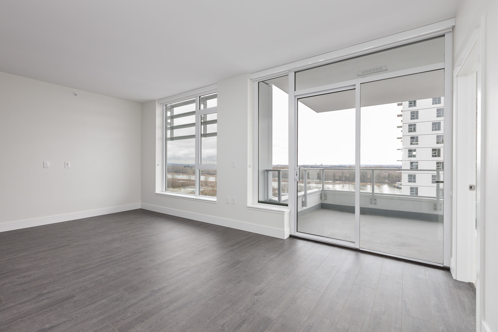 Empty modern apartment with large windows, sliding glass door leading to a balcony, and dark wood flooring.